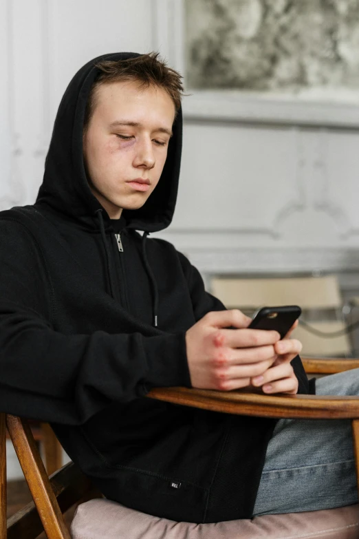 a man sitting in a chair looking at his cell phone, trending on pexels, hyperrealism, wearing a black hoodie, 1 7 - year - old boy thin face, 15081959 21121991 01012000 4k, incompetent