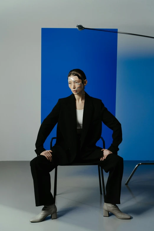 a man sitting on a chair in front of a blue backdrop, inspired by Marina Abramović, photograph of a techwear woman, wearing wool suit, humanoid, girl with pearl earring