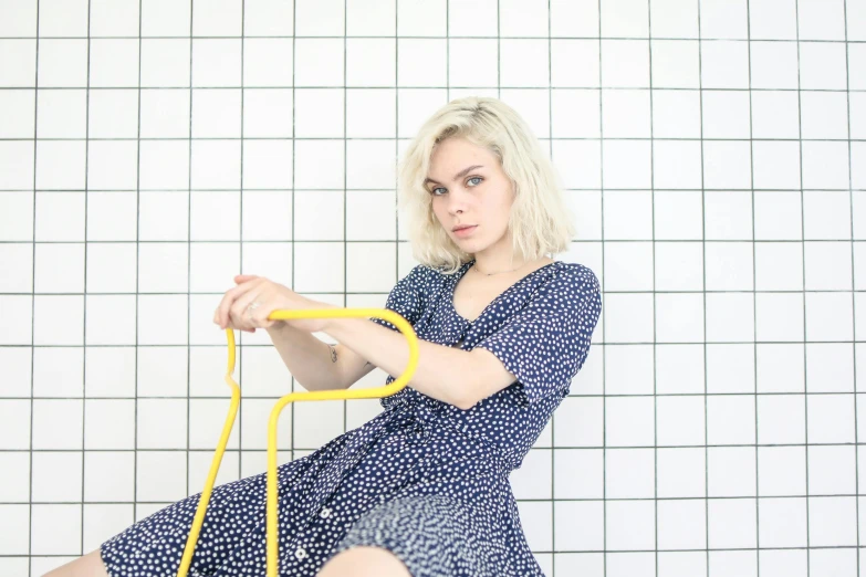 a woman sitting on a stool holding a yellow object, inspired by Louisa Matthíasdóttir, pexels contest winner, blue checkerboard dress, young blonde woman, charli xcx, polka dot