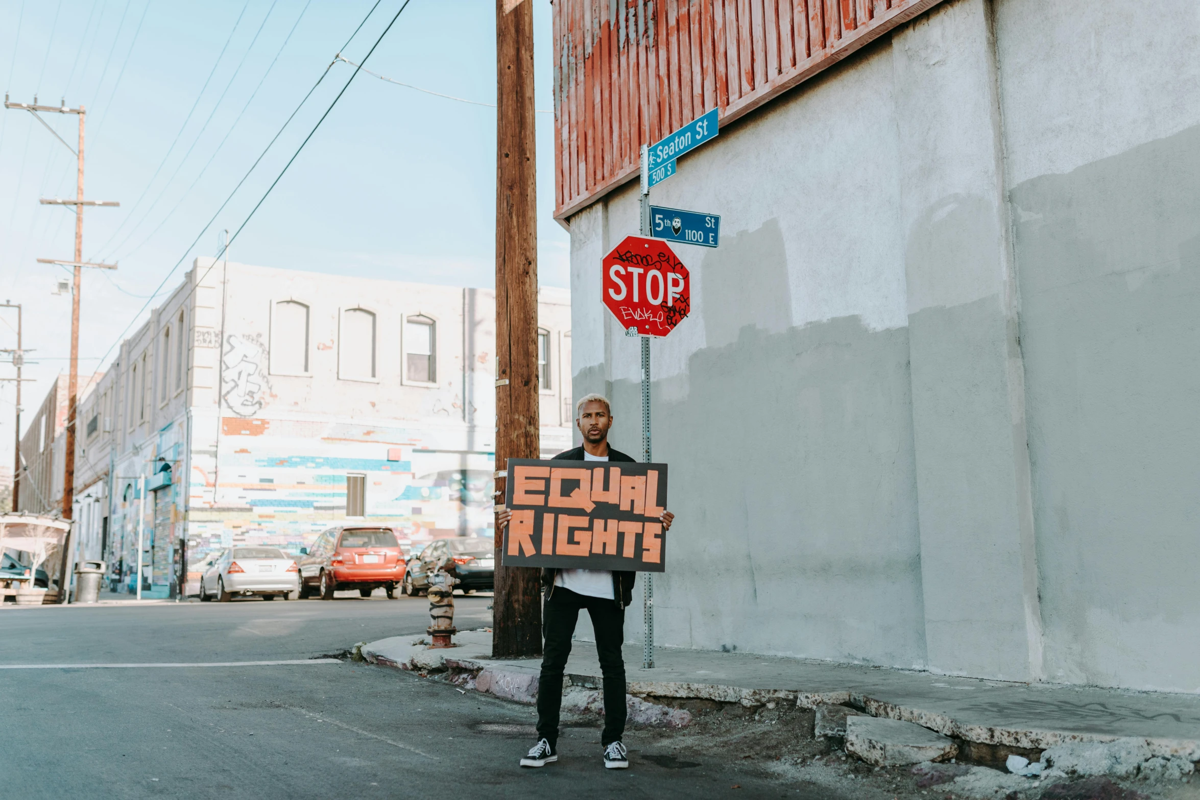 a man holding a sign on the side of a street, by Julia Pishtar, pexels contest winner, black arts movement, lgbtq, background image, los angeles ca, standing in township street