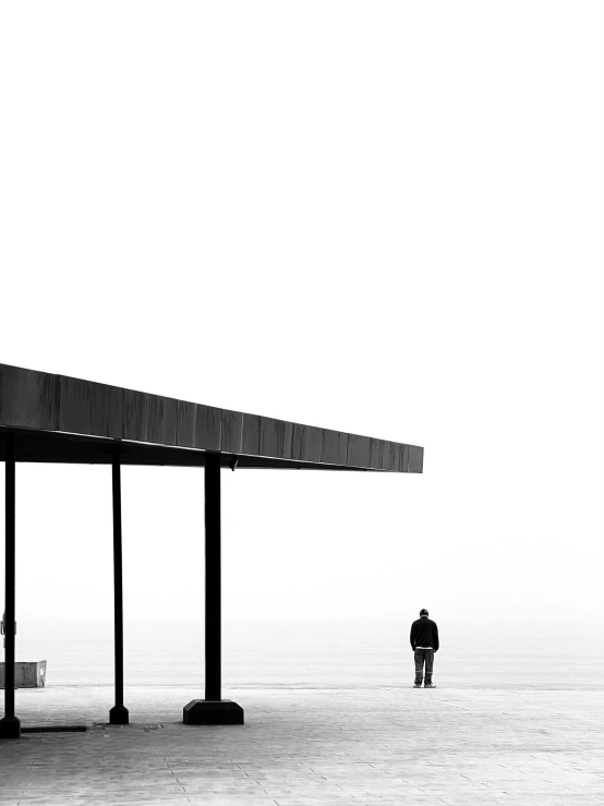 a black and white photo of a person standing in front of a building, by Zsolt Bodoni, unsplash contest winner, postminimalism, next to the sea, bus stop, pavilion, sittin