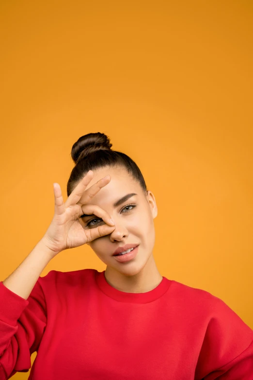 a woman covering her eyes with her hands, trending on pexels, in front of an orange background, tight bun, isabela moner, pointing