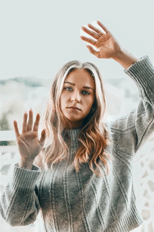 a woman standing in front of a window with her hands in the air, inspired by Louisa Matthíasdóttir, trending on pexels, grey sweater, sydney sweeney, head shot, hand gesture