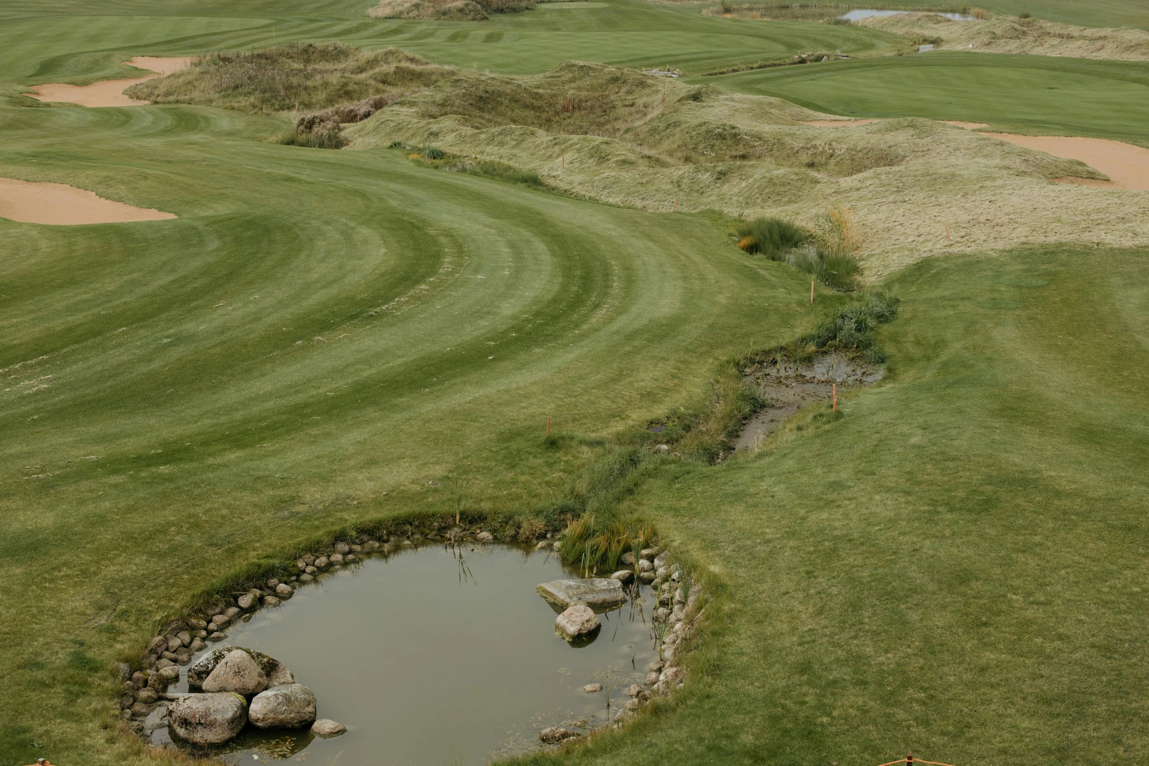 a golf course with a pond in the middle, by David Simpson, unsplash, land art, erosion channels river, whealan, grassy hill, thumbnail