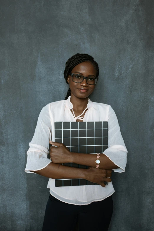 a woman standing in front of a wall holding a solar panel, by Chinwe Chukwuogo-Roy, renaissance, wearing square glasses, grey backdrop, holding notebook, high-quality photo