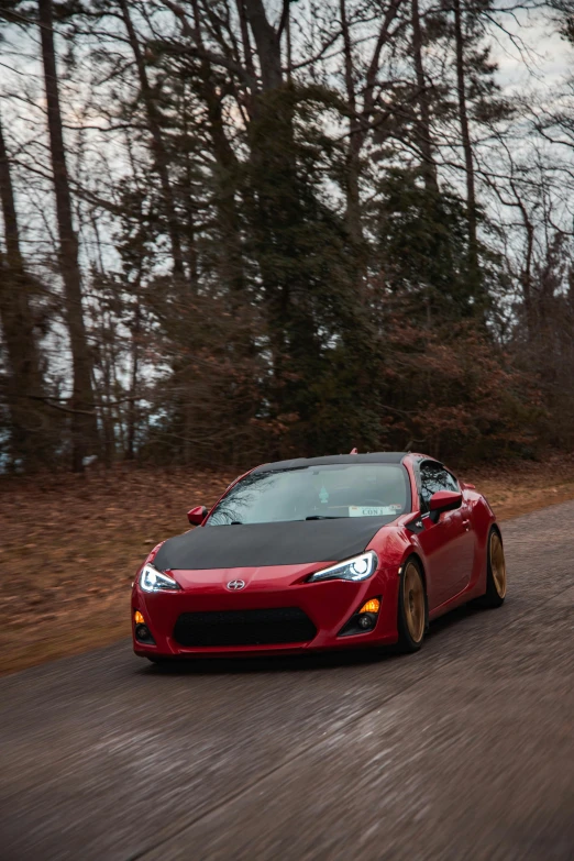 a red car driving down a road next to a forest, samurai vinyl wrap, golden detailing, front flash, panorama shot