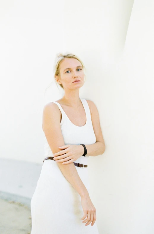a woman in a white dress leaning against a wall, by Sara Saftleven, minimalism, wearing a tanktop, confident expression, white building, a blond