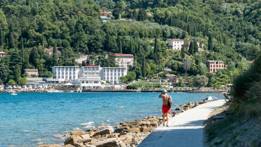 a man walking down a path next to a body of water, a picture, croatian coastline, multiple stories, onsens, commercially ready