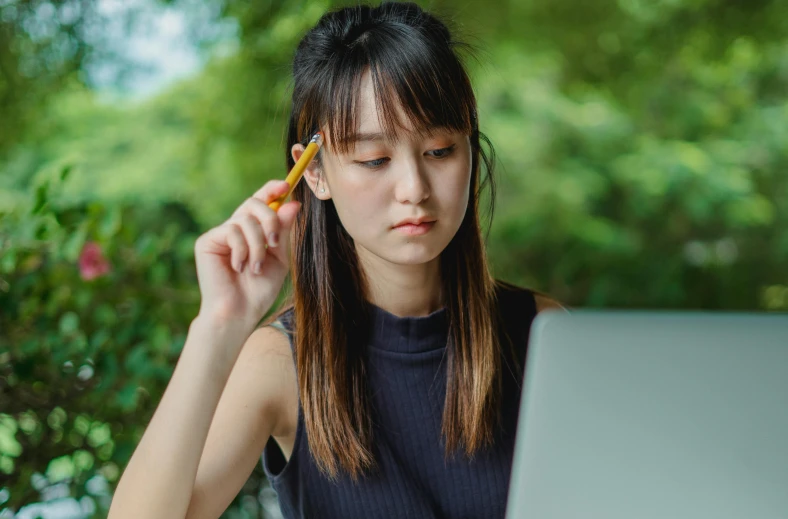 a woman sitting in front of a laptop with a pencil in her hand, trending on pexels, asian face, bare forehead, student, performing