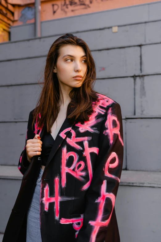 a woman standing in front of a wall with graffiti on it, an album cover, by Julia Pishtar, trending on pexels, wearing a blazer, hot pink and black, brutalist fashion show, portrait sophie mudd