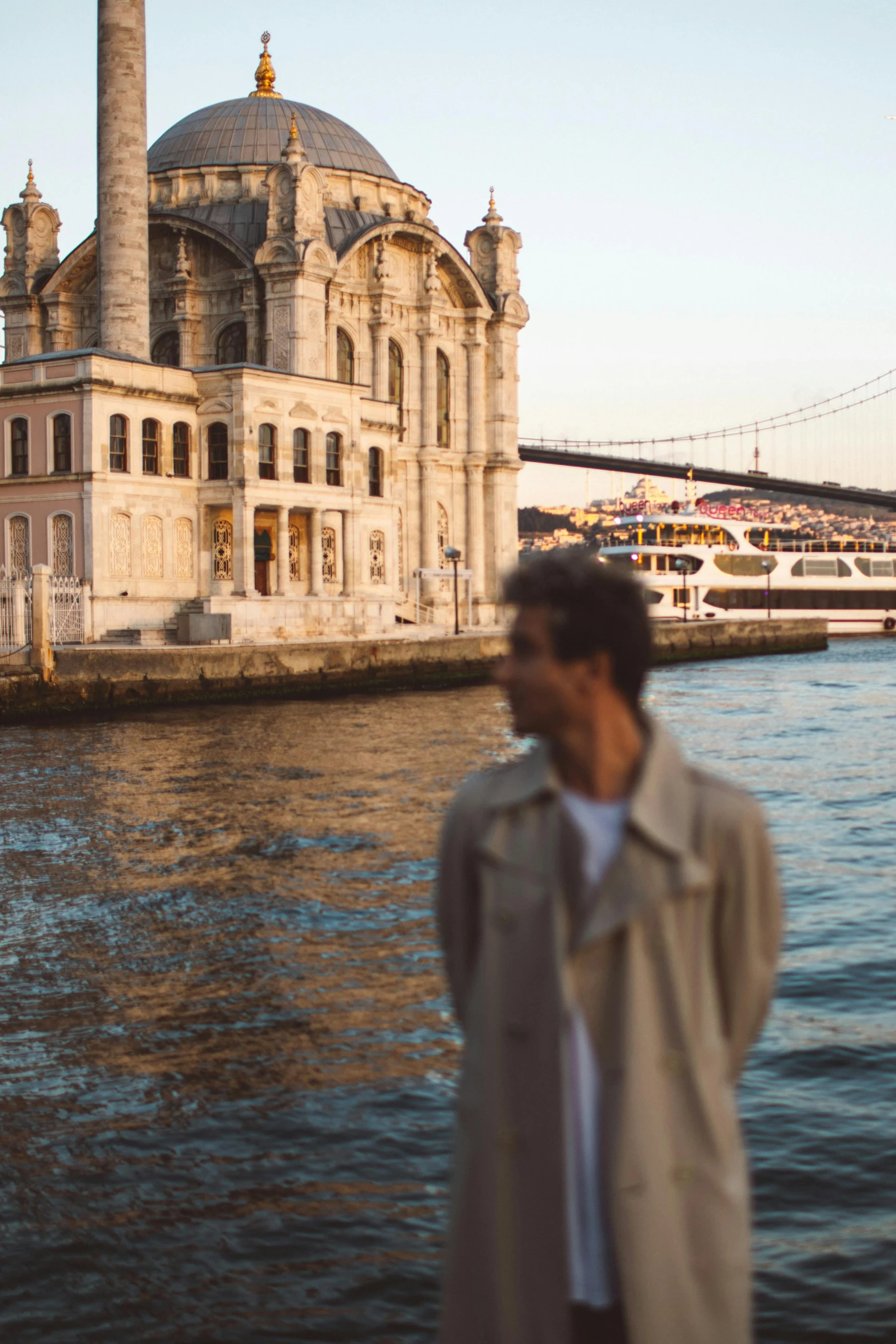 a man standing next to a body of water, a picture, inspired by Nadim Karam, pexels contest winner, renaissance, architecture in the background, light brown trenchcoat, istanbul, zayn malik