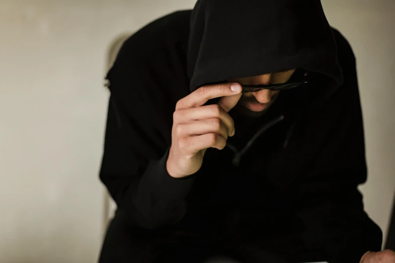 a person in a black hoodie using a cell phone, shy looking down, wearing black glasses, shroud, sitting on a table