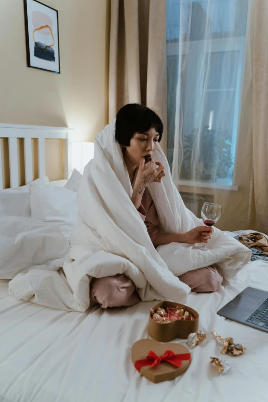 a woman sitting on top of a bed next to a laptop, drinking wine, tech robes, coughing, having a snack