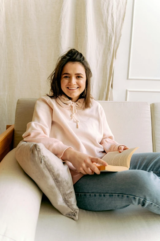 a woman sitting on a couch reading a book, a portrait, pexels contest winner, wearing a pastel pink hoodie, maisie williams, wearing a linen shirt, happily smiling at the camera