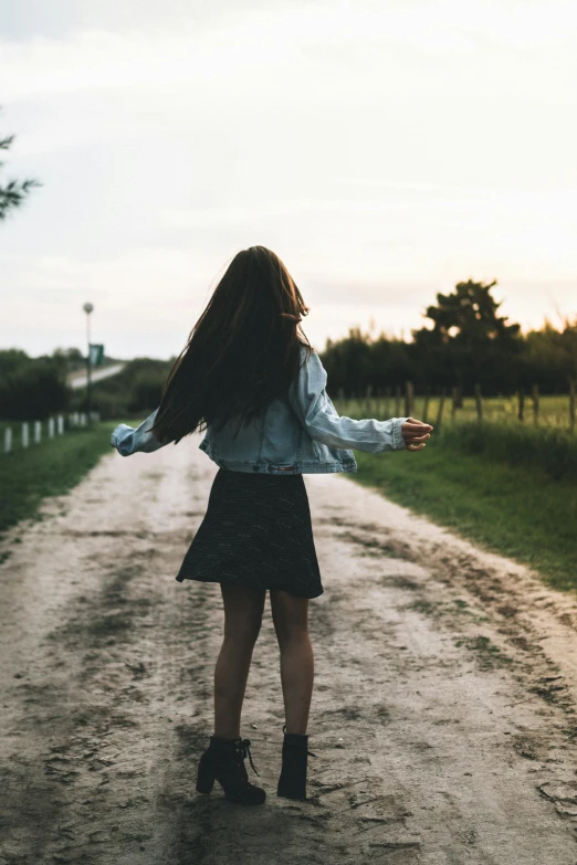 a woman standing on a dirt road with her arms outstretched, pexels contest winner, renaissance, girl with dark brown hair, wearing skirt, walking away, teen girl