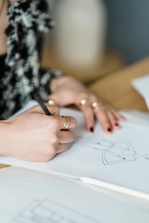 a woman sitting at a table writing on a piece of paper, a drawing, wearing a designer top, gold details, 9 9 designs, zoomed in