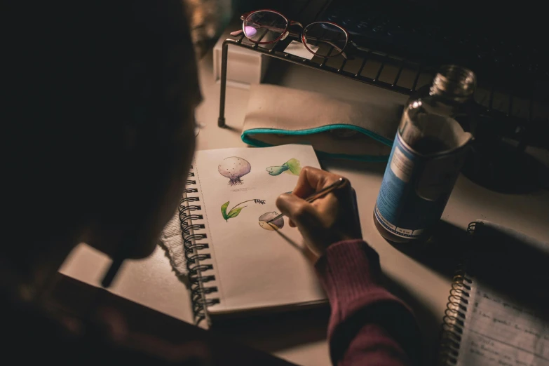 a person sitting at a desk drawing on a piece of paper, a drawing, by Olivia Peguero, pexels contest winner, light watercolour, the lighting is dark, a high angle shot, student