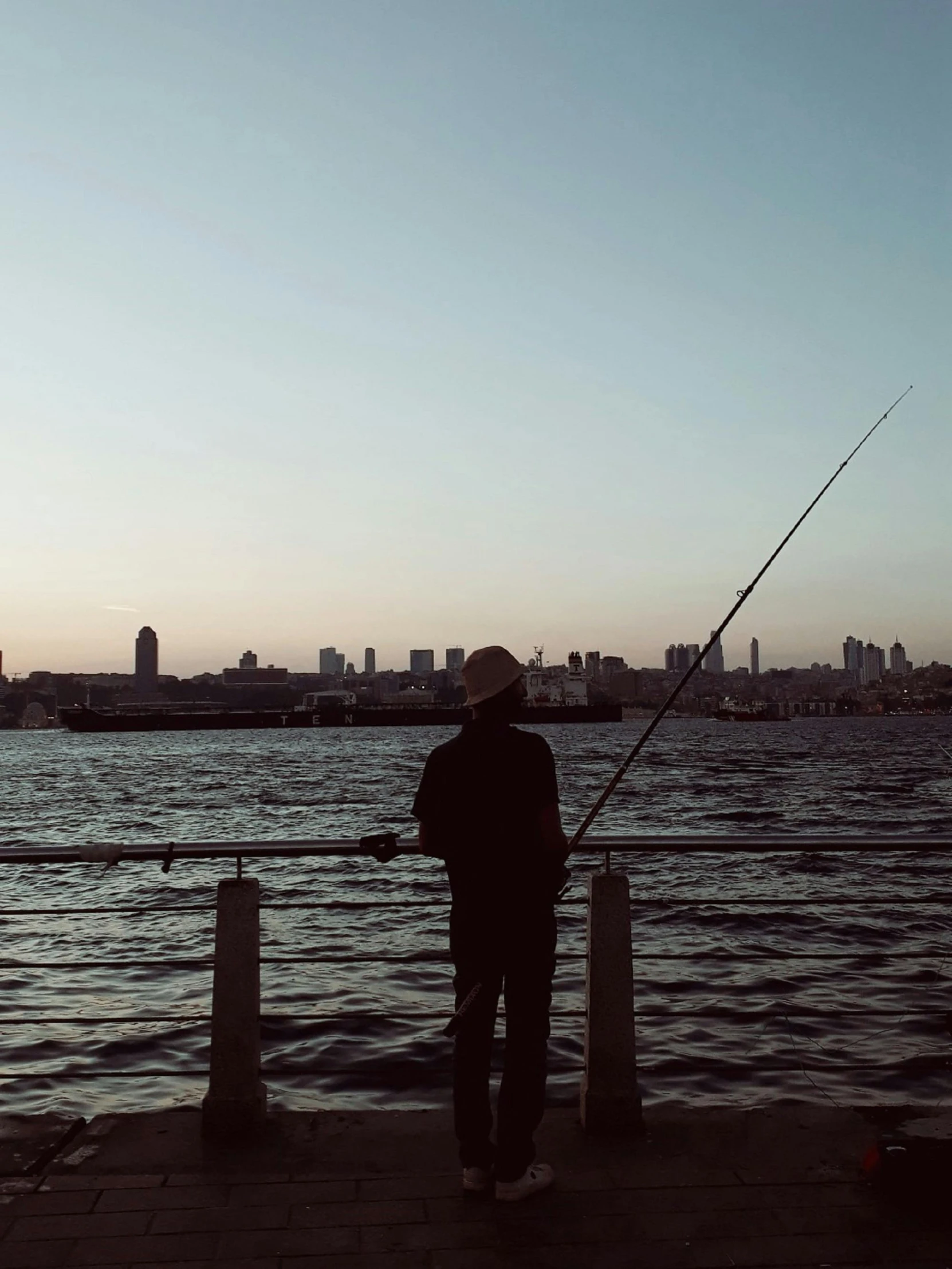 a man standing on top of a pier next to a body of water, fishing pole, golden hour in tokyo, watching new york, lo-fi