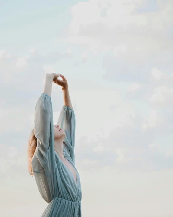 a woman standing on top of a sandy beach, aestheticism, raising an arm, pale blue skin, wearing a grey robe, unsplash transparent