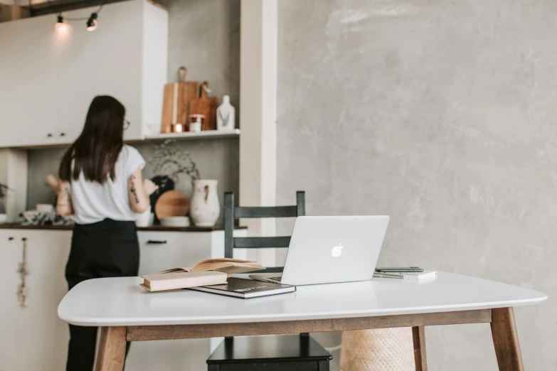 a woman standing in a kitchen next to a table with a laptop on it, by Nicolette Macnamara, trending on pexels, minimalist desk, background image, profile image, wooden desks with books