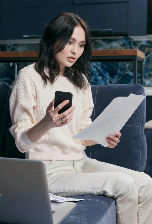 a woman sitting on a couch holding a cell phone, papers on table, jackie tsai style, smart casual, promo image