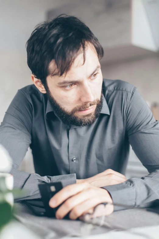 a man sitting at a table looking at his cell phone, beard stubble, in gunmetal grey, pensive, someone lost job