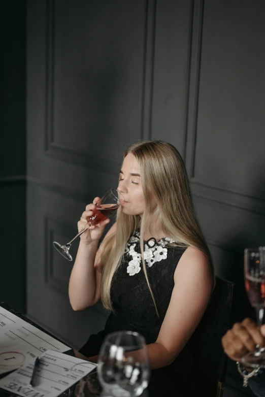 a woman sitting at a table with a glass of wine, bubbly, a girl with blonde hair, dark backdrop, licking out