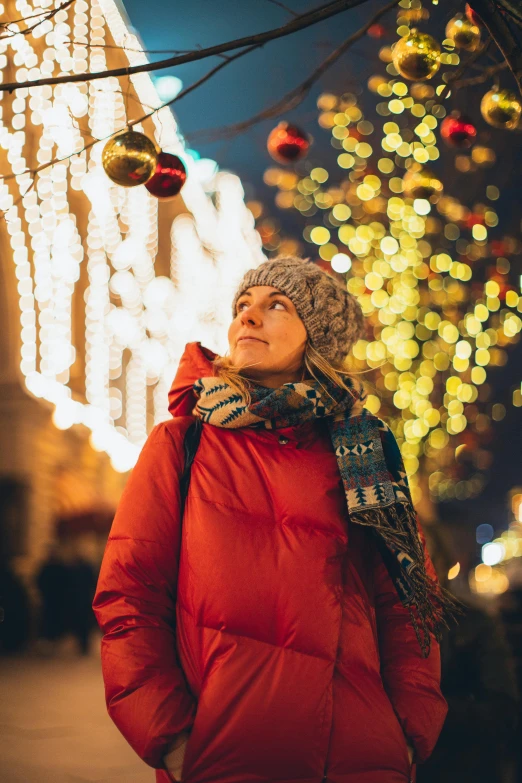 a woman standing in front of a christmas tree, bright city lights, warm color clothes, looking upward, promo image