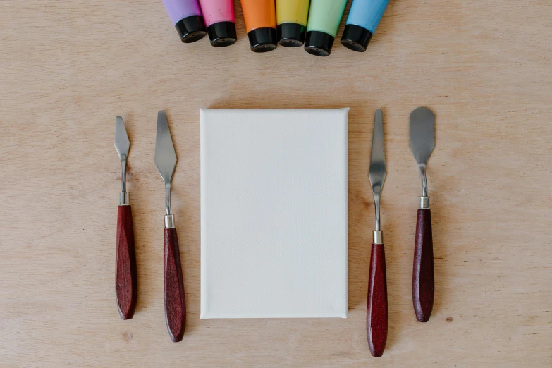 a book sitting on top of a wooden table next to a knife and fork, a minimalist painting, inspired by Kyffin Williams, pexels contest winner, multicoloured, miniature product photo, unfinished canvas, white