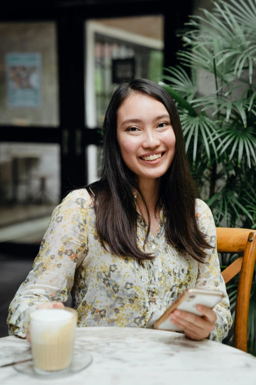 a woman sitting at a table with a glass of milk, joy ang, long sleeves, south east asian with round face, brown