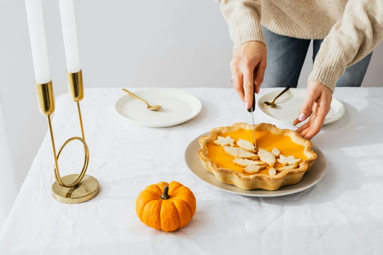 a woman decorating a pumpkin pie on a table, by Nicolette Macnamara, natural candle lighting, set against a white background, cutlery, on a candle holder