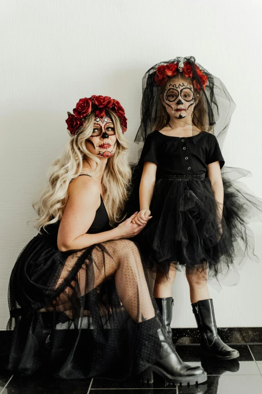 a couple of women sitting next to each other, an album cover, pexels contest winner, lowbrow, dia de muertos dress and make up, with a kid, she is wearing a black dress, 2019 trending photo