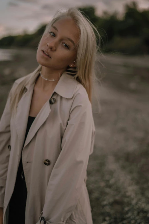 a woman standing on a beach next to a body of water, an album cover, trending on pexels, aestheticism, light brown trenchcoat, white - blond hair, 🤤 girl portrait, low lights