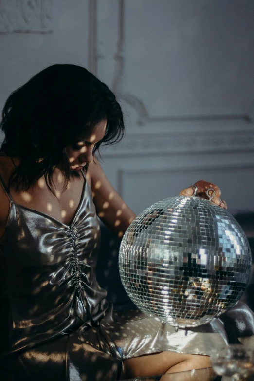 a woman sitting on a couch holding a disco ball, inspired by Elsa Bleda, fancy silver dress, admiring her own reflection, 7 0 s vibe, standing in a dimly lit room