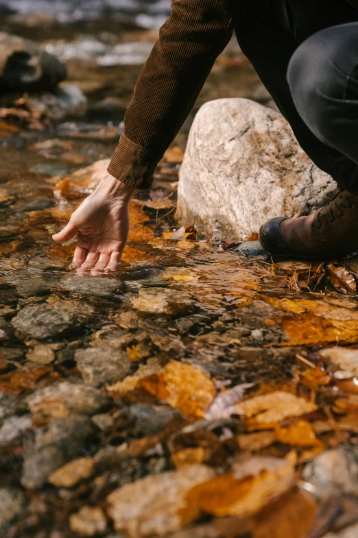 a man kneeling on top of a rock next to a river, by Jessie Algie, pexels contest winner, symbolism, flowing clear water creek bed, touches of gold leaf, detail shot, autumnal