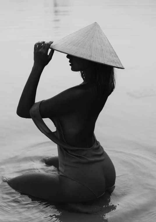 a woman sitting in the water with a hat on her head, a black and white photo, inspired by Herb Ritts, unsplash contest winner, in style of lam manh, perfect female body silhouette, bum, hooded
