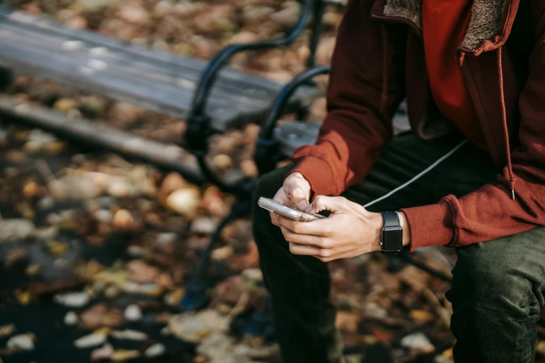 a man sitting on a bench looking at his cell phone, by Emma Andijewska, trending on pexels, happening, red shirt brown pants, wearing a watch, autumnal, close up to the screen