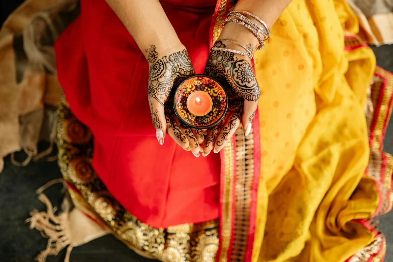 a woman holding a lit candle in her hands, trending on pexels, hurufiyya, wearing traditional garb, black and yellow and red scheme, indian ink, flatlay