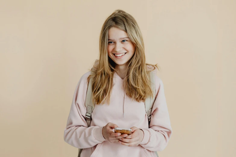 a girl in a pink sweatshirt holding a cell phone, by Emma Andijewska, trending on pexels, smiling girl, beige hoodie, portrait of high school girl, plain background