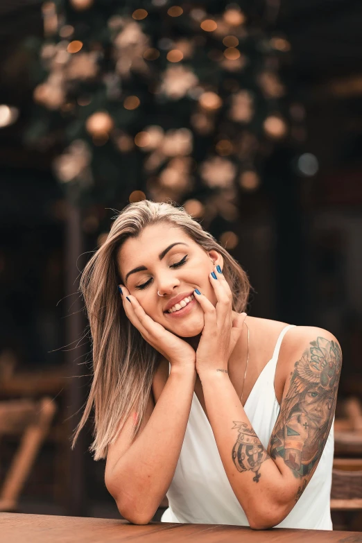 a woman sitting at a table with her hand on her face, a tattoo, trending on pexels, happy fashion model face, a gorgeous blonde, dazzling lights, 2019 trending photo