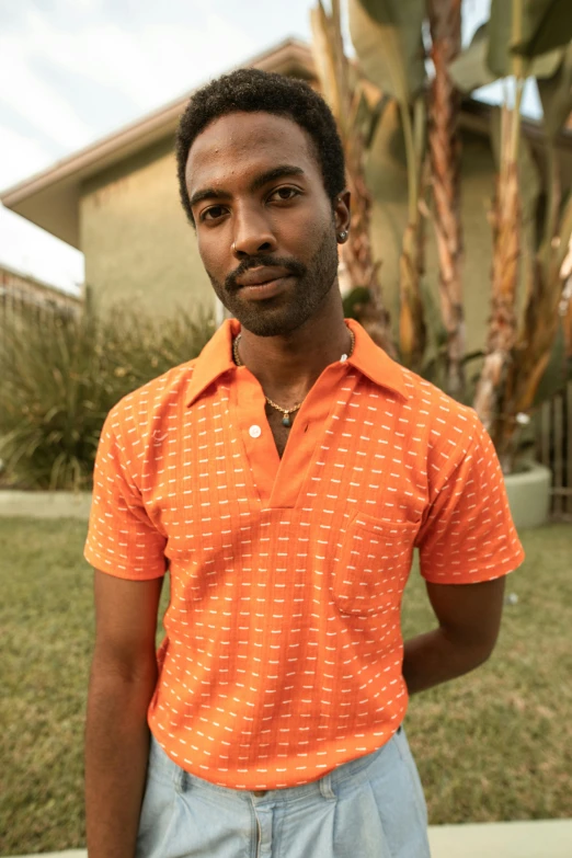 a man standing in front of a palm tree, an album cover, wearing an orange t shirt, wearing polo shirt, seventies cinestill, thumbnail