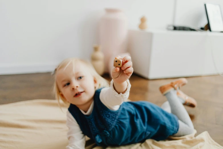 a little girl laying on the floor holding a cookie, by Emma Andijewska, pexels contest winner, wooden art toys, pointing at the camera, gif, holding a bell