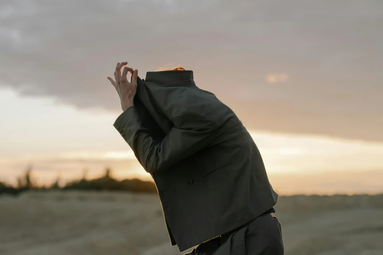 a man in a suit standing in a field, an album cover, by Emma Andijewska, unsplash, surrealism, cloak covering face, bending down slightly, in the sunset, wearing a worn out suit