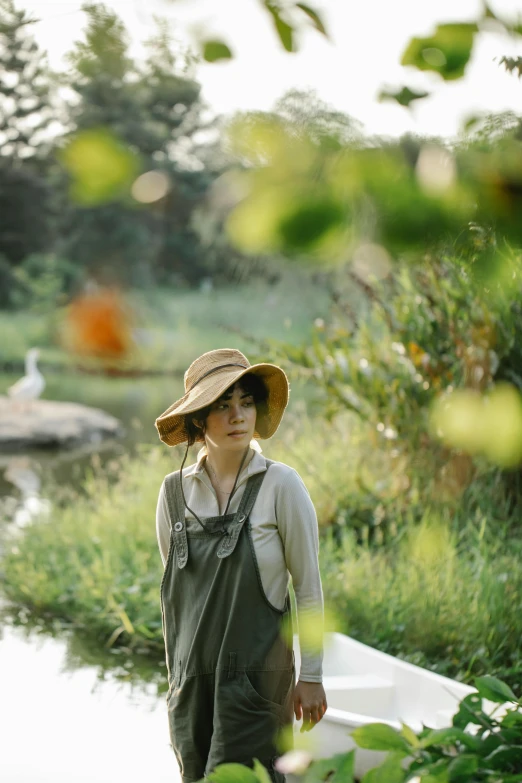 a woman in a hat standing next to a body of water, inspired by Jung Park, unsplash, renaissance, wearing farm clothes, in a verdant garden, wearing overalls, sangyeob park