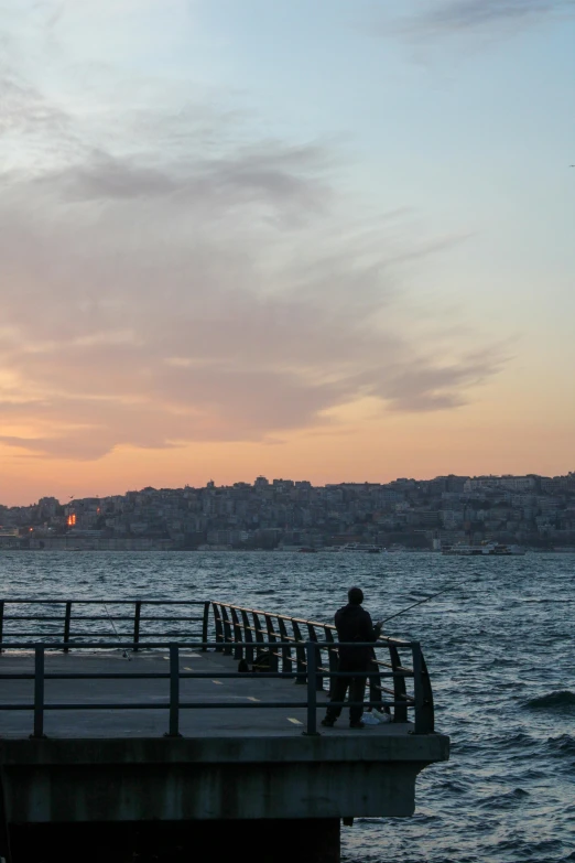 a man standing on top of a pier next to the ocean, istanbul, sunset in the distance, manly, city views
