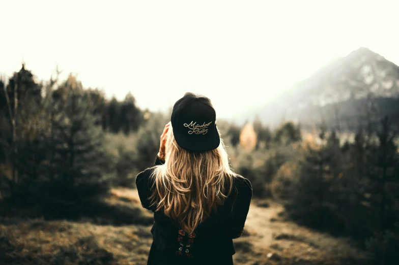 a woman standing in a field talking on a cell phone, a picture, by Emma Andijewska, trending on pexels, realism, wearing a backwards baseball cap, pinterest hair picture, on a mountain, embroidered velvet