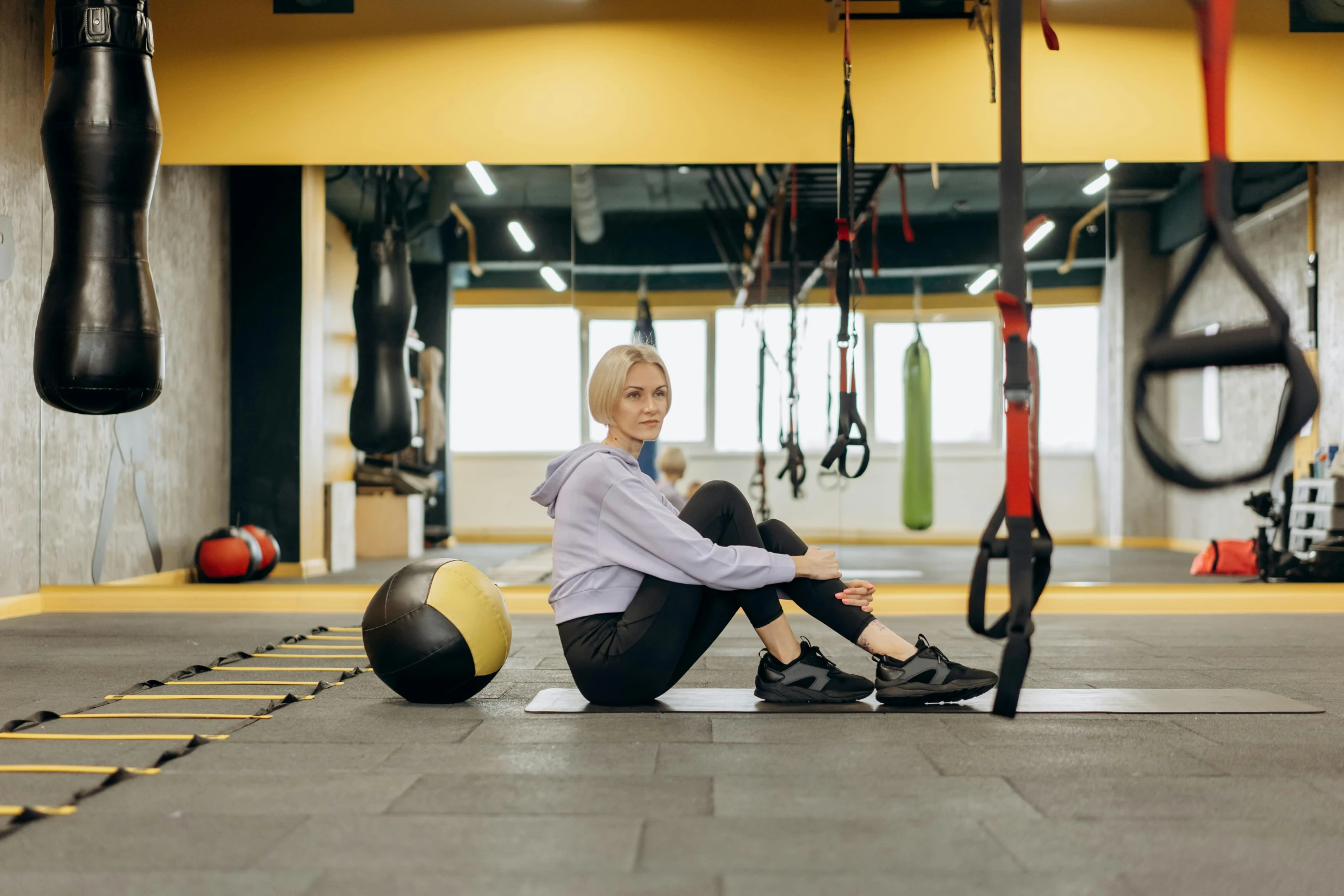 a woman sitting on the ground with a medicine ball, by Emma Andijewska, pexels contest winner, hurufiyya, background a gym, avatar image, yellow carpeted, distant photo