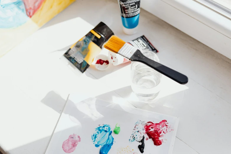 a white table topped with art supplies next to a window, inspired by artist, pexels contest winner, melting paint, holding a paintbrush, on a canva, jpeg artefacts on canvas