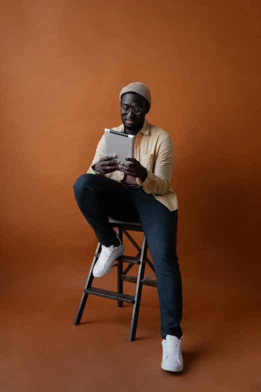 a man sitting on a stool holding a tablet, by Carey Morris, pexels contest winner, ( brown skin ), plain background, librarian, jaylen brown