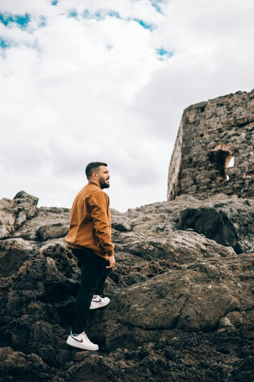 a man standing on top of a rocky hill, by Jesper Knudsen, happening, walking in a castle, streetwear, brown jacket, man with beard
